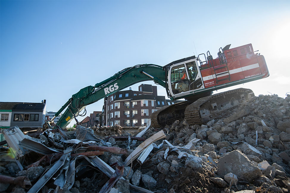 Graafmachine van KGS verricht sloopwerkzaamheden op een berg puin tijdens stedelijk infraproject met woningen op de achtergrond met een open hydraulisch systeem